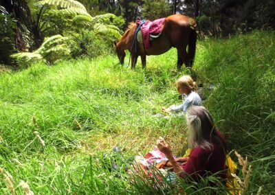 girl and old lady sitting in tall grass having a picnic while their brown horse is grazing right by their side