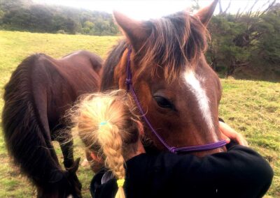 girl in black top and with blond braid intimately hugging the nose of an unhlatered dark brown horse with white stripe and black mane, a second horse in background close by