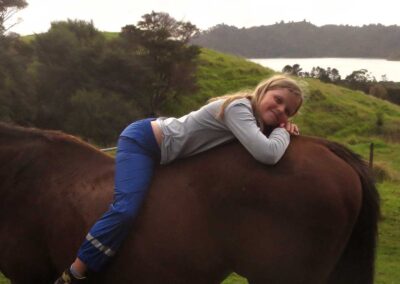 young smiling girl in rainpants lying on her tummy on the back of a chestnut horse