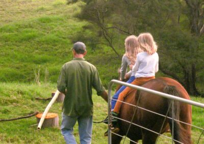 rear view of man in olive green shirt, jeans and black cap leading chestnut horse with two girls in white tops and with long blond hair sitting bareback on the horse