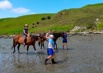 4 peaople and 2 horses walking over the empty tidal zone of an estuary, grass covered hills behind them, one person sitting bareback on one of the horses, all people wearing hats