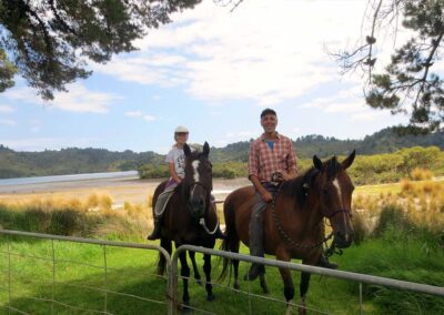 2 smiling riders – girl and man – and their horses by a closed gate on lush grass in front of estuary