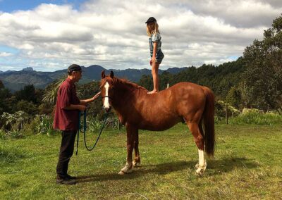 man in red shirt and black pants and cap holding chestnut horse on slack lead rope, horse with white stripe on nose and white feet, girl wearing shots and a cap standing up straight and barefoot on horse's back, dramatic landscape and sky in background