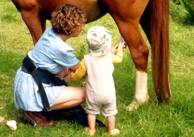 kneeling mother in blue dress and standing baby child with hat looking at a horse's hind leg, holding a comb