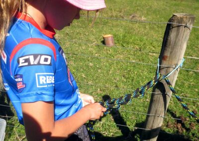 girl in blue sports teeshirt and hat tying a horse knot to a fence post