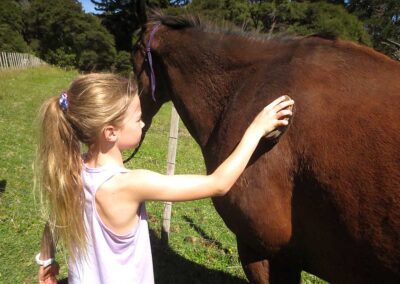 young blond girl with pony tail brushing the shoulder of a brown horse