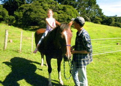 little girl sitting bareback on standing chestnut horse with white stripe on forehead, man in dark blue checkered shirt and black cap holding slack leadrope and looking at girl, large hilly paddock and forested area in background