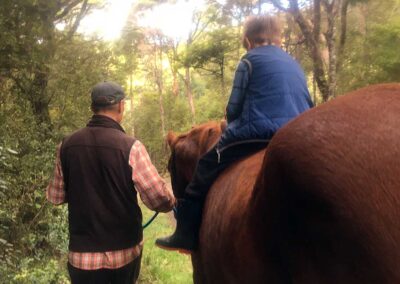 man in shirt and black vest and cap leading chestnut horse with little boy riding bareback through NZ bush